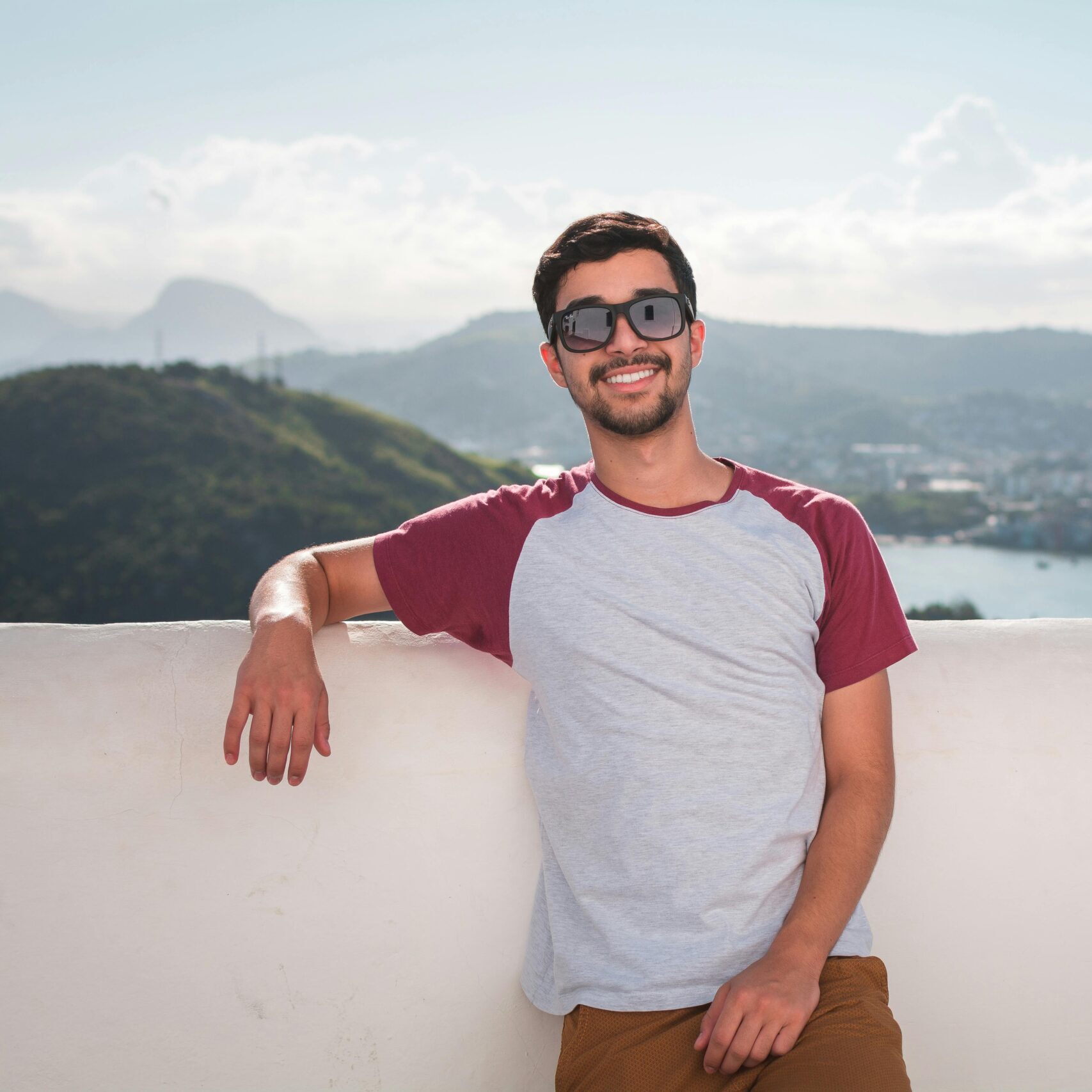 Young man in sunglasses leaning on a balcony with a scenic city view in the background.