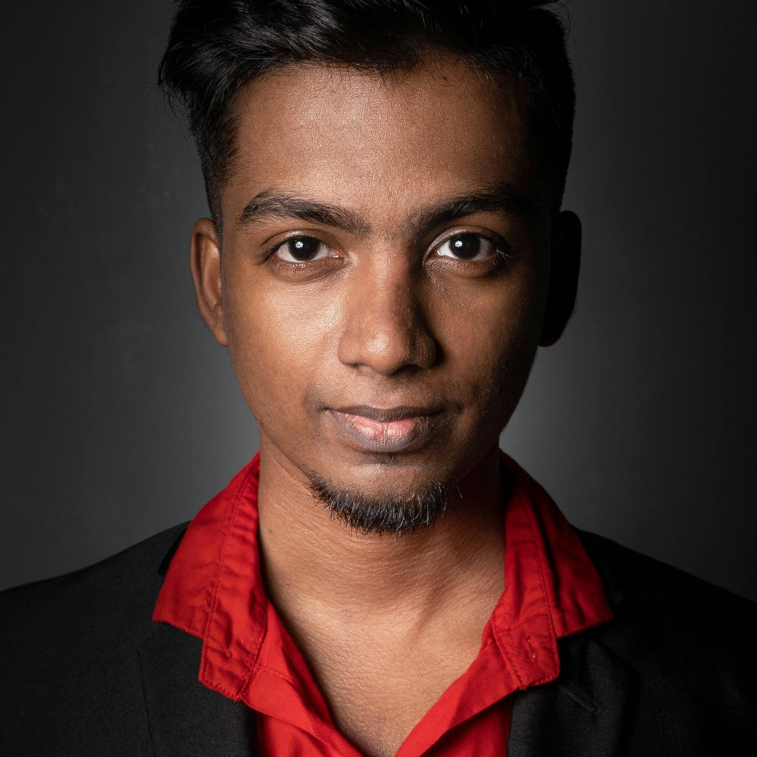 Close-up portrait of a young man in a black blazer and red shirt, smiling at the camera.