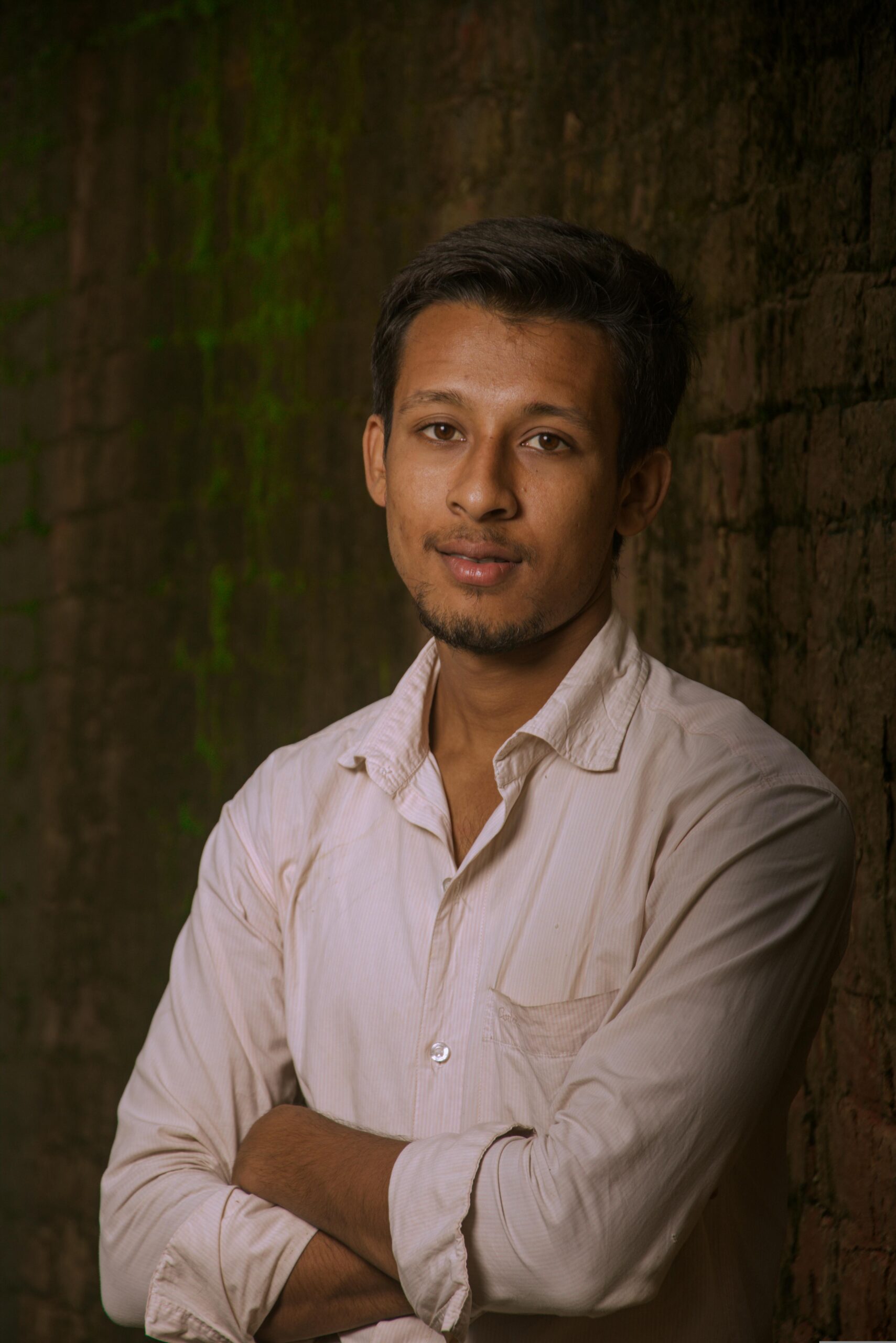 Portrait of a young man in India, exuding confidence with a subtle smile against a rustic brick wall.